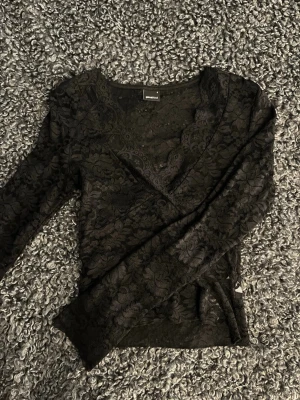 Svart spetsblus med omlott - Snygg svart blus i spets med omlottdesign och v-ringning. Blusen har långa ärmar och ett blommigt spetsmönster som ger en elegant look. Perfekt för dig som vill ha något extra till din outfit.