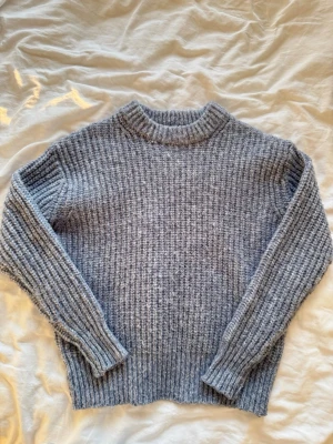 Grå ribbstickad tröja - Mysig blå ribbstickad tröja med rund hals och långa ärmar. Tröjan har en chunky struktur och breda muddar vid ärmslut och nederkant. Perfekt för kalla dagar och enkel att matcha med olika outfits.