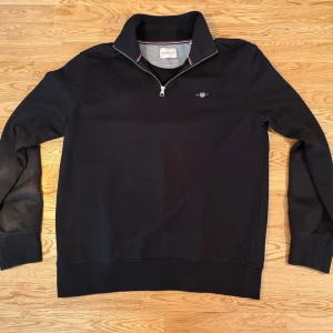 Svart halvzip tröja från Gant - Klassisk svart halvzip tröja från Gant med broderad logga på bröstet. Tröjan har hög krage med dragkedja och diskreta ränder i rött och vitt på insidan av kragen. Perfekt för en stilren och avslappnad look.