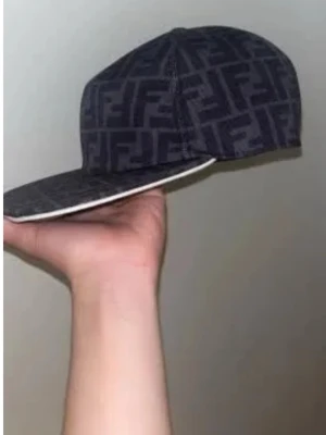 Svart keps - Säljer en svart keps med fin FF-mönster över hela kepsen. Kepsen har rak skärm och klassisk snapback-form. Materialet ser ut att vara bomull eller en bomullsblandning. Perfekt för dig som gillar streetwear och vill sticka ut.
