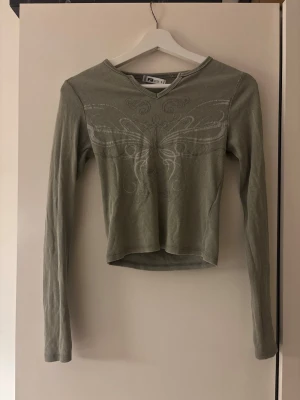 Grön ribbad långärmad topp - Snygg grön ribbad topp med v-ringning och lång ärm. Toppen har ett vitt tryck framtill i form av ett abstrakt mönster och är croppad i modellen. Perfekt att styla med jeans eller kjol för en trendig look.