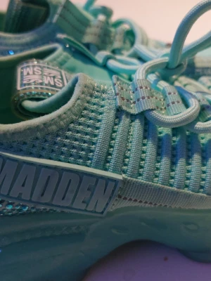 Turkosa sneakers från Steve Madden - Snygga turkosa sneakers från Steve Madden med stickad ovandel och coola detaljer. Skorna har elastiska snören och tydlig branding på sidan. Sulan är rejäl och matchar färgen på ovandelen. Perfekta för dig som vill sticka ut med färg och stil.