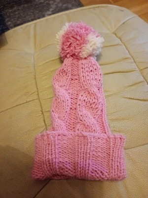Rosa stickad mössa med tofs - Söt rosa stickad mössa med kabelstickat mönster och en fluffig tofs i rosa och vitt upptill. Mössan har vikt kant och är gjord i mjukt garn, perfekt för kyliga dagar.material  akryl.