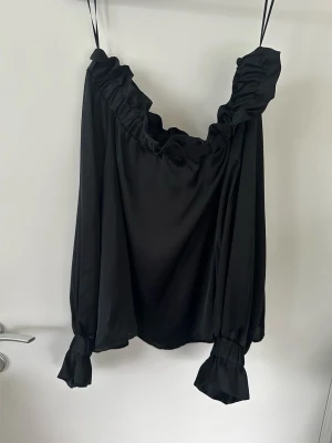 Svart offshoulder blus med volang -storlek M, Gina Tricot  - Härlig svart axelbandslös / off-shoulder blus / topp i satin från Gina Tricot i storlek M. Fin axelbandslös modell med långa ärmar och smockad ringning och ärmslut. Superlent halvglansigt satintyg. Mycket bra skick, som ny!