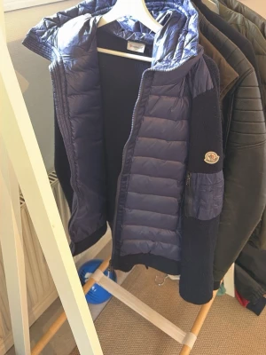 Mörkblå cardigan från Moncler - Snygg mörkblå dunjacka från Moncler med quiltad front och huva. Ärmarna är stickade i matchande blått och jackan har en dragkedja framtill. Moncler-logga på vänster ärm. Perfekt för kalla vinterdagar.