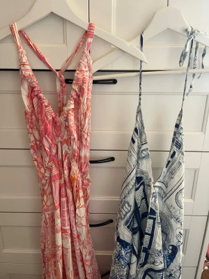 Långklänning med grafiskt mönster - Två maxiklänningar med grafiskt mönster, en i rosa och orange toner och en i blått och vitt. Båda har smala axelband, v-ringad hals och lös passform. Perfekta för sommaren med sitt lätta bomullsmaterial och snygga detaljer. STORLEKEN ÄR ONE SIZE