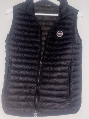 Svart dunväst från Colmar - Snygg svart dunväst från Colmar med quiltad design och två dragkedjeförsedda fickor. Västen har hög krage och Colmar-logga på bröstet. Perfekt för lager-på-lager och ger en sportig look. Materialet är lätt och smidigt.
