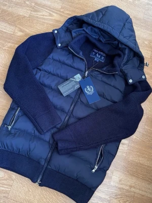 Mörkblå dunväst med huva - Snygg mörkblå dunväst från Royal Air Force med avtagbar huva och stickade ärmar. Västen har två dragkedjeförsedda fickor och ribbade muddar. Perfekt för kalla vinterdagar och ger en sportig look.