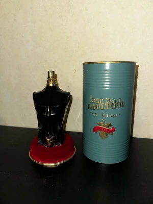 Jean Paul Gaultier Le Beau Parfum - Snygg parfymflaska formad som en svart manlig torso med gulddetaljer från Jean Paul Gaultier. Kommer med originalförpackning i blått och guld. Flaskan står på en röd sammetsliknande bas. Perfekt för dig som gillar unika och lyxiga dofter.