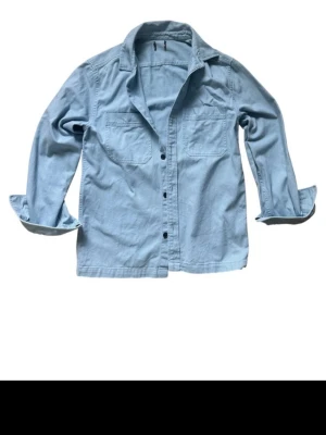 Ljusblå overshirt - Snygg ljusblå overshirt med två bröstfickor och knäppning framtill. Skjortan har en avslappnad passform och långa ärmar som kan vikas upp för en mer casual stil. Perfekt att bära över en t-shirt eller skjorta. Modellen är 183 och väger 70kg. Lappen är borta så vet inte märke.