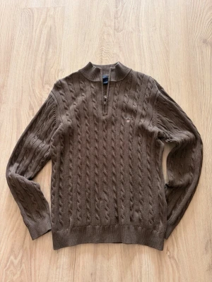 Brun kabelstickad tröja från GANT - Snygg brun kabelstickad tröja från GANT med halv dragkedja och ribbad krage. Tröjan har långa ärmar, diskret GANT-logga på bröstet och är tillverkad i mjukt material. Perfekt för lager på lager och en klassisk look.