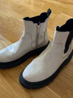 Ljusbeige chunky Chelsea boots från Zara - Säljer ett par ljusbeige Chelsea boots från Zara med grov svart sula och dragkedja på sidan. Bootsens ovandel är i glansigt material och har elastiska partier på sidorna för enkel påtagning. Perfekta för dig som vill ha en edgy och trendig look. Använt men gott skick, små sprickor i ovandelen där foten böjs, se bild 4! 