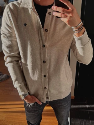 Beige cardigan från Morris (Aldrig använd) - Snygg beige cardigan från Morris med knappar framtill och liten broderad logga på bröstet. Tröjan har krage, ribbade muddar och är tillverkad i mjukt material som passar perfekt till lager på lager. Enkel att matcha med olika outfits.