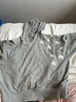 Grå hoodie med vita stjärnor - Mysig grå hoodie med flera vita stjärnor på framsidan och en stor vit stjärna på ryggen. Hoodien har en klassisk huva och ribbade muddar nedtill. Perfekt för dig som gillar en enkel men ändå unik look.