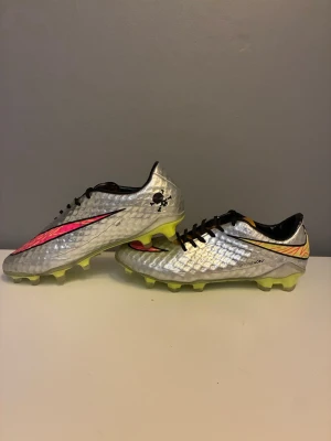 Silvriga Nike Hypervenom fotbollsskor - Priset kan definitivt diskuteras Säljer ett par Nike Hypervenom fotbollsskor i silver med svarta och rosa detaljer samt neongul sula. Skorna har låg profil, snörning och mönstrad ovandel för bättre bollkontroll. Perfekta för dig som vill sticka ut på planen.