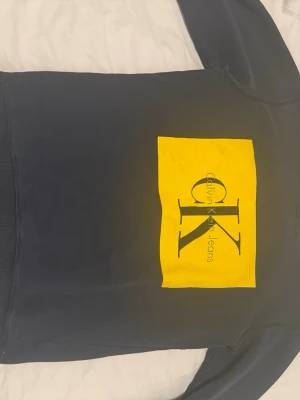 Mörkblå calvin klein tröja - Snygg svart hoodie från Calvin Klein Jeans med stor gul rektangel och CK-logga på bröstet. Klassisk passform med långa ärmar och ribbade muddar. Perfekt för dig som gillar streetwear och vill sticka ut med en tydlig logga. Medium storlek men passar S också 