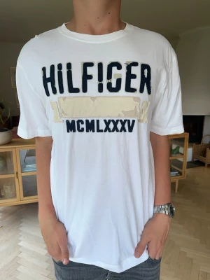 Hilfiger T-shirt vintage herr vit med tryck - Snygg vit t-shirt från Tommy Hilfiger med maffigt broderat Hilfiger-tryck och romerska siffror (MCMLXXXV) på bröstet. Ger en skön, sportig retrolook. Väldigt fint skick och skönt material. Rent och fräscht från djurfritt hem.#tommyhilfiger #hilfigertshirt #mcmlxxxv #vittshirt #retro #streetwear #casual #märkeskläder #herr #vinted