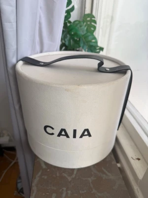  CAIA julkalender box - Säljer en vit rund box från CAIA med svart handtag och tydlig logga framtill. Väskan är tillverkad i ett kraftigt tygmaterial och har en stilren, minimalistisk design. Perfekt för dig som gillar unika accessoarer och vill sticka ut.