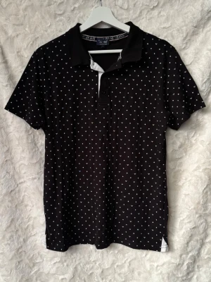 Armani jeans Svart t-shirt storlek XL - Svart pikétröja från Armani Jeans med diskret AJ-mönster över hela tröjan. Klassisk pikékrage, kort ärm och knappslå framtill. Passar både till jeans och en mer dressad outfit.