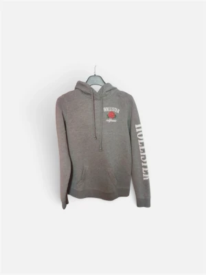 Grå hoodie från Hollister med tryck - Snygg grå hoodie från Hollister med vit logga och röd ros på bröstet samt stort vitt tryck på ena ärmen. Klassisk känguruficka framtill och justerbar huva med dragsko. Perfekt för en avslappnad och trendig look.