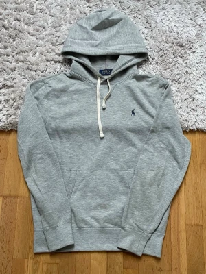 Grå hoodie från Polo Ralph Lauren - Snygg grå hoodie från Polo Ralph Lauren med klassisk huva och dragsko. Har en liten mörkblå logga broderad på bröstet och en stor magficka framtill. Tillverkad i mjukt bomullsmaterial, perfekt för chill dagar eller när du vill ha en clean look.