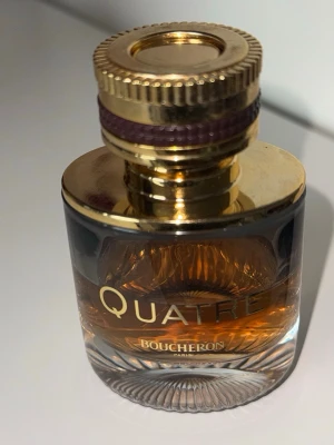Boucheron Quatre parfym 50ml - Elegant parfymflaska från Boucheron, modell Quatre 50ml, lite mer än hälften kvar. Flaskan är rundad med tjockt glas och har en lyxig guldfärgad kork med lila detalj. Vätskan inuti är bärnstensfärgad och flaskan har tydliga guldiga logotyper. Perfekt för dig som gillar exklusiva dofter.