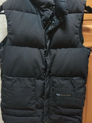 Svart dunväst med hög krage - Snygg svart dunväst med hög krage och tryckknappar framtill. Västen har två stora framfickor och en diskret logga på ena fickan. Perfekt för lager-på-lager och håller dig varm under kyliga dagar. Canada gosse väst