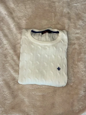 Vit kabelstickad tröja - Kabelstickad vit tröja med rund halsringning och diskret broderad logga i mörkblått på bröstet. Tröjan har långa ärmar och ribbade muddar vid ärmslut och nederkant. Perfekt för en clean och klassisk stil.