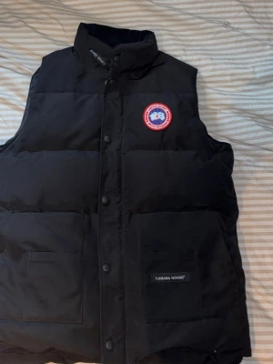 Canada goose väst - Säljer en svart dunväst från Canada Goose med klassisk logga på bröstet och två stora fickor framtill. Västen har hög krage och stängs med tryckknappar. Perfekt för kalla dagar när du vill hålla värmen och ändå se stilren ut.