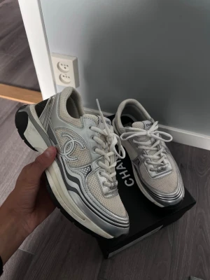 Chanel skor - Snygga vita sneakers från Chanel med silverfärgade detaljer och tydlig logga på sidan. Skorna har en chunky sula i vitt och svart, mesh- och läderpaneler samt klassisk snörning. Perfekta för dig som vill ha en exklusiv och trendig look.