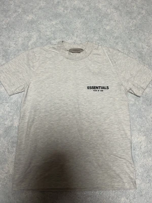 Ljusgrå Essentials T-shirt Fear of God - Snygg ljusgrå T-shirt från Essentials Fear of God med svart logga på bröstet. Klassisk rund hals och korta ärmar. Perfekt för en clean och stilren look. Tillverkad i mjuk bomull för maximal komfort.