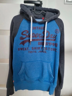 Blå och grå hoodie från Superdry - Snygg hoodie från Superdry i blått med mörkgrå ärmar och huva. Stor logga och text framtill i vinrött. Mjuk insida, känguruficka och vita dragsnören i huvan. Perfekt för en avslappnad streetstyle-look.