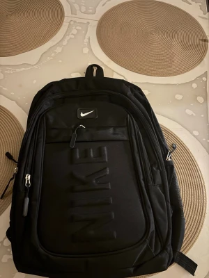 Svart ryggsäck från Nike - Snygg svart ryggsäck från Nike med flera dragkedjefack och vadderade axelband. Stor Nike-logga på framsidan och diskret Swoosh-märke upptill. Tillverkad i slitstarkt material och har praktiska sidofickor.