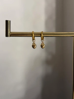 Guldiga örhängen med hjärtberlocker - Säljer ett par stilrena örhängen i guldfärgad metall med små hjärtformade berlocker. Örhängena har en enkel och modern design som passar till många olika stilar. Perfekta för dig som gillar minimalistiska smycken med en söt twist.