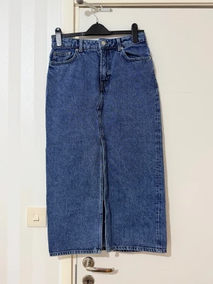 Lång jeanskjol med slits framtill - Säljer en klassisk blå lång jeanskjol i rak modell med hög midja och slits framtill. Kjolen har fem fickor, bälteshällor och stängs med knapp och dragkedja. Perfekt till vardags för en avslappnad men trendig look.