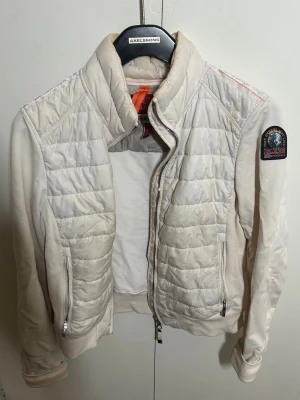 Vit quiltad jacka från Parajumpers - Snygg vit quiltad jacka från Parajumpers med hög krage och dragkedja framtill. Jackan har ett emblem på ärmen och två fickor med dragkedja. Materialmix med vadderade partier och mjuka ärmar ger en sportig look.