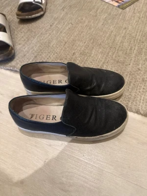 Svarta slip-on sneakers Tiger of Sweden - Säljer ett par svarta slip-on sneakers från Tiger of Sweden. Skorna har en vit tjock sula och ovandel i svart texturerat material. De är låga och har en enkel, stilren design utan snörning. Perfekta för en clean och avslappnad look.