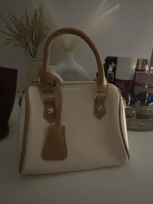 Vit och beige handväska med detaljer - Snygg handväska i vitt med beigea detaljer och handtag. Väskan har en strukturerad yta, guldfärgade metalldetaljer och dragkedja upptill. Perfekt storlek för det viktigaste och en stilren accessoar till din outfit.