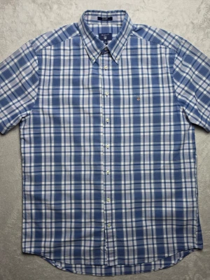 Gant Tech Prep Regular Fit kortärmad skjorta rutig blå/vit storlek L / 41/42 - Kortärmad rutig skjorta från Gant i Tech Prep-serien och Regular Fit. Klassisk blå och vit design med diskreta röda detaljer, button-down-krage och broderad Gant-logotyp på bröstet. En lättburen modell som passar bra till både vardag och mer uppklädda sommartillfällen.  Storlek L / 41/42 Regular Fit Tech Prep  Mycket fint skick.  Mått Bröstbredd: ca 57 cm Plagglängd: ca 75 cm Axelbredd: ca 47 cm  Material 78% bomull 22% polyester