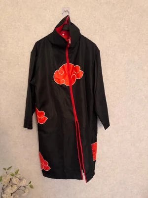Akatsuki cosplaykappa med huva - Säljer en svart cosplaykappa inspirerad av Akatsuki från Naruto. Kappan har röda moln med vit kant, röd dragkedja och huva. Perfekt för cosplay eller maskerad. Materialet är tunt tyg, troligen polyester.