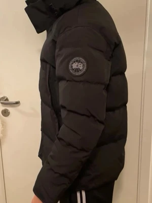 Canada goose Wyndham  - Säljer en svart pufferjacka med quiltad design och hög krage. Jackan har dragkedja framtill och en patch på ärmen. Perfekt för kalla dagar under höst och vinter. Snygg och varm modell med normal passform.
