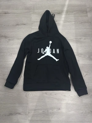 Svart Jordan hoodie  - Snygg svart hoodie från Jordan med den klassiska Jumpman-loggan och texten 'JORDAN' i vitt på bröstet. Hoodien har huva och en stor magficka framtill. Perfekt för en sportig och avslappnad stil.
