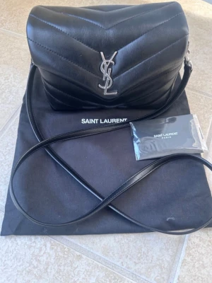 Svart Saint Laurent handväska i läder - Snygg svart handväska från Saint Laurent i quiltat läder med silverfärgad YSL-logga framtill. Väskan har en lång axelrem och klassisk, stilren design. Perfekt för dig som gillar lyxiga accessoarer och tidlös stil.