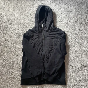 Svart cardigan  - Svart cardigan med quiltad front och huva. Tröjan har dragkedja hela vägen och en liten gul logga på ärmen. perfekt för kyliga dagar. Snygg och enkel design som funkar till det mesta. Skriv om du har en fråga. Ny pris runt 1500kr