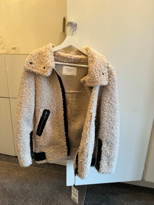 Beige teddyjacka med svarta detaljer - Säljer en beige teddyjacka med stora slag och svarta dragkedjor vid fickorna. Jackan har en oversized look och är gjord i mjukt, fluffigt material som ger en riktigt cozy vibe. Perfekt för kyliga dagar när du vill vara både varm och stylish.