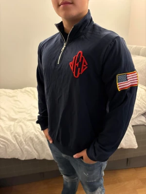 Mörkblå hoodie med USA-flagga - Snygg mörkblå hoodie med halv dragkedja och hög krage. På bröstet finns ett rött broderat märke och på ena ärmen sitter en patch med amerikanska flaggan. Hoodien har en avslappnad passform och är tillverkad i mjukt material, perfekt till jeans. Passar folk mellan 175-185, Den är jätte mörkblå