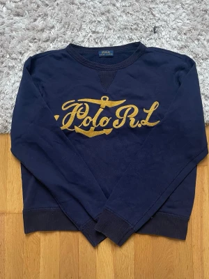 Marinblå sweatshirt Polo Ralph Lauren - Mörkblå sweatshirt från Polo Ralph Lauren med gul broderad text och ankare på bröstet. Tröjan har rund halsringning, ribbade muddar vid ärmslut och nederkant. Perfekt för en avslappnad och stilren look.