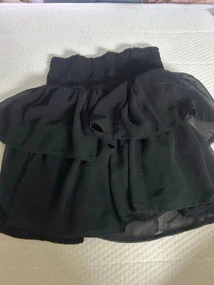 Två volangkjolar i svart och vitt - Säljer två korta kjolar med volanger, en i svart och en i vitt. Båda har bred smock i midjan och är gjorda i ett tunt, flowy material som ger en luftig känsla. Perfekta för sommarens alla tillfällen!
