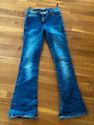 Blå bootcut jeans med slitningar - Säljer ett par blå bootcut jeans med snygga slitningar och tvättade detaljer. Jeansen har klassisk femficksmodell, dragkedja och knapp i midjan. Dom är ganska små i storleken . Perfekta för dig som gillar retrovibbar och utsvängda ben.