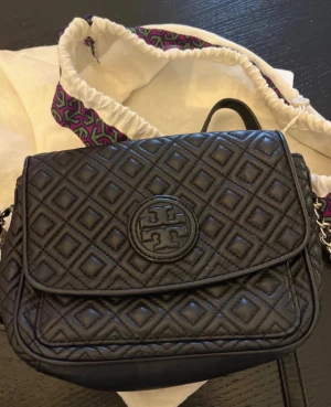 ÄKTA Tory Burch quiltad handväska väska - Snygg svart quiltad handväska från Tory Burch med rutmönster och rund logga på locket. Väskan har en kedjerem med läderdetaljer och ett lock som stängs framtill. Har äkthetsverifierats hos Vestaire Collective.